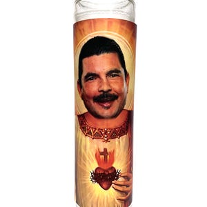 Puede incluir: Una vela de vidrio transparente con una etiqueta impresa que presenta el retrato de un hombre, un corazón con llamas e imágenes religiosas. La vela tiene un interior de cera blanca y está diseñada para uso decorativo o espiritual.