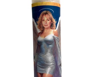 May include: A clear glass candle with a label featuring an image of a woman with angel wings. The label reads "Saint Samantha" in a brown font. The candle is filled with white wax.