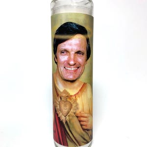 May include: A clear glass prayer candle featuring a portrait of a smiling man with a halo. The image is set against a light background, with the man wearing a gold and red garment and a gold heart emblem.