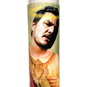 May include: A clear glass prayer candle featuring an image of a man with a halo and a gold heart emblem. The man has dark hair and a beard, and is dressed in religious garb. The candle is a novelty item.