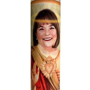May include: A clear glass candle featuring a portrait of a smiling woman, with a halo and heart-shaped emblem. The candle has a red and gold colour scheme, creating a religious or devotional theme.