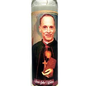 Op de afbeelding: Een heldere glazen kaars met een portret van Saint John Waters. De afbeelding toont een man in een zwarte toga die een boek en een kruis vasthoudt. De kaars heeft een witte was binnenkant en een label met "Saint John Waters".