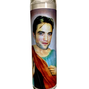 May include: A clear glass candle with a portrait of a man in a saintly pose. The man has light skin, dark hair, and is wearing a blue tunic and a red drape. The candle is filled with white wax.