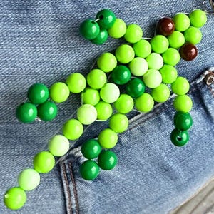 Op de afbeelding: Een kralen hagedis figuur gemaakt van groene kralen in verschillende tinten, met bruine kralen voor de ogen. De kralen zijn geregen op draad, waardoor een eigenzinnig, handgemaakt ontwerp ontstaat. De hagedis wordt getoond tegen een denim achtergrond.