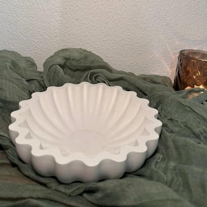 May include: A white, scalloped-edge bowl with a textured surface. The bowl has a fluted interior design and sits on a draped, olive-green fabric. A candle in a brown glass holder is in the background.