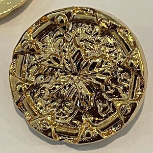 Könnte beinhalten: Nahaufnahme eines goldfarbenen, runden Knopfes mit einem verzierten, strukturierten Design. Der Knopf weist ein erhabenes, detailliertes Muster mit einem zentralen floralen Motiv und geometrischen Bordüren auf. Der Knopf ist wahrscheinlich aus Metall und hat ein glänzendes Finish.
