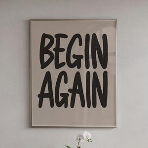 Può includere: Una stampa incorniciata con le parole "BEGIN AGAIN" in grassetto nero. Il testo è centrato su uno sfondo beige chiaro. La cornice è sottile e color argento. Un fiore bianco è visibile in basso.