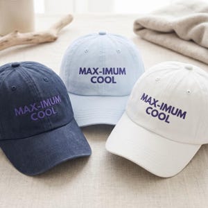 Op de afbeelding: Drie baseballpetten in marineblauw, lichtblauw en wit. Elke pet is voorzien van de tekst MAX-IMUM COOL in donkerpaars borduursel op het voorpaneel. De petten hebben een gestructureerde kroon, een gebogen klep en een knoopje bovenop.