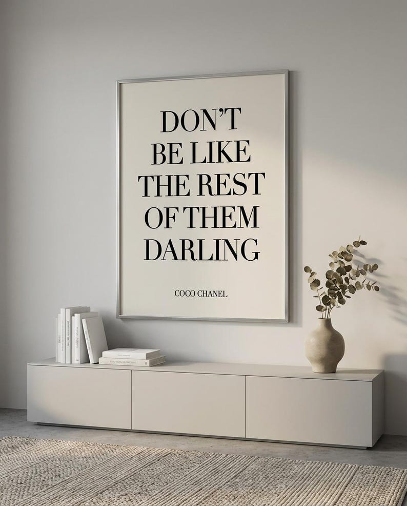 Puede incluir: Un cuadro enmarcado con la frase "DON'T BE LIKE THE REST OF THEM DARLING" atribuida a Coco Chanel. El cuadro est&aacute; sobre un mueble claro con libros y un jarr&oacute;n con plantas secas.