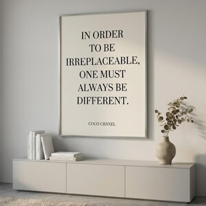 Puede incluir: Un cuadro enmarcado con la cita "IN ORDER TO BE IRREPLACEABLE, ONE MUST ALWAYS BE DIFFERENT." de Coco Chanel. El cuadro est&aacute; sobre un mueble gris claro con libros y un jarr&oacute;n.