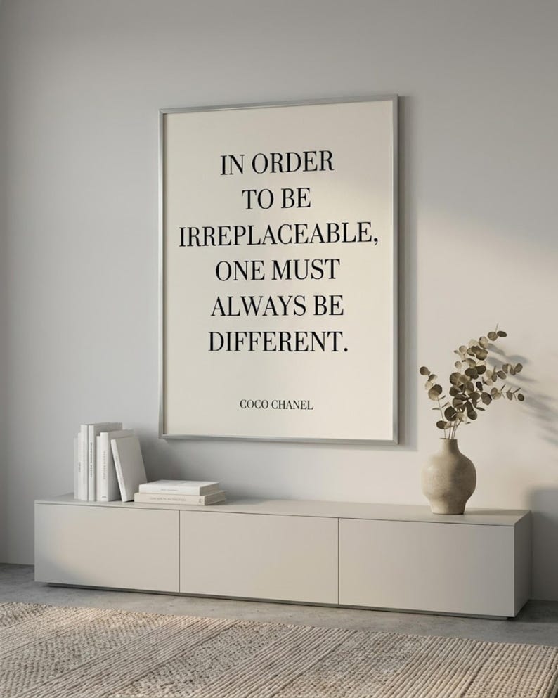Puede incluir: Un cuadro enmarcado con la cita "IN ORDER TO BE IRREPLACEABLE, ONE MUST ALWAYS BE DIFFERENT." de Coco Chanel. El cuadro est&aacute; sobre un mueble gris claro con libros y un jarr&oacute;n.