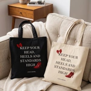 Könnte beinhalten: Zwei Tragetaschen, eine schwarze und eine cremefarbene, mit dem Text "KEEP YOUR HEAD, HEELS AND STANDARDS HIGH" und einer roten High-Heel-Grafik. Die Taschen liegen auf einem beigen Sofa mit einer weißen Decke.