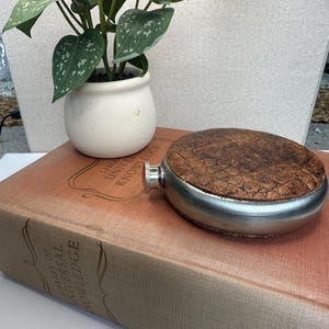 May include: A stainless steel hip flask with a brown, textured leather top. The flask sits on a vintage book with the words "Library of Universal Knowledge". A small potted plant is in the background. The book is in the foreground.