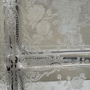 May include: Close-up of a silver-toned decorative tray with an embossed floral and scroll design. The intricate pattern features roses and other flowers, with detailed borders and elegant curves. The tray's surface has a reflective quality.