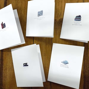 May include: Five white greeting cards with watercolour illustrations of books. Each card has a different image of books and a quote. The cards are arranged on a wooden surface.