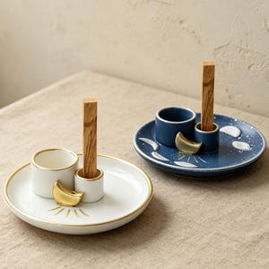 Puede incluir: Dos porta-inciensos de cerámica, uno blanco y otro azul oscuro, cada uno con una bandeja circular, una pequeña taza y un soporte cilíndrico que sostiene un palito de madera. Ambas bandejas tienen motivos dorados de media luna y rayos de sol.