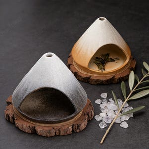 May include: Two ceramic incense holders with a conical shape, each set on a wooden coaster. One is gray and the other is cream and orange. The holders have a large opening on the side and a small hole on top. The cream and orange holder contains dried herbs.