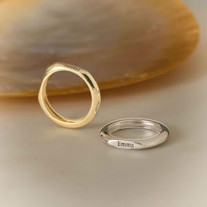 Könnte beinhalten: Ein goldener Ring mit der Gravur Hanna und ein silberner Ring mit der Gravur Emma. Beide Ringe haben ein glattes, abgerundetes Banddesign. Die Ringe liegen nah beieinander und zeigen ihre metallischen Oberflächen sowie die deutliche Beschriftung.