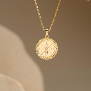 May include: A gold-toned necklace featuring a circular pendant with a textured, beaded border. The center of the pendant is divided into four quadrants by intersecting lines, each containing the engraved letters A, J, S, and N in a cross formation.