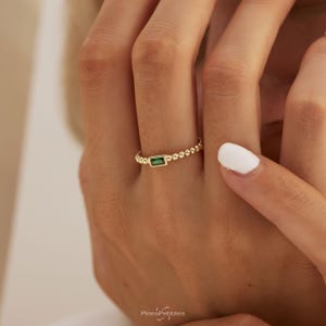 Könnte beinhalten: Ein goldener Ring mit einem kleinen, rechteckigen grünen Edelstein in der Mitte. Die Ringschiene besteht aus einer Reihe kleiner, miteinander verbundener goldener Kugeln. Der Text PinesPebbles ist am unteren Bildrand sichtbar.