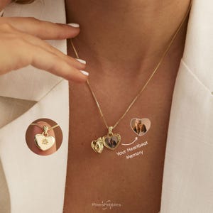 Op de afbeelding: Een gouden hartvormige medaillonketting aan een fijne ketting. Het medaillon kan worden geopend en toont een kleine foto aan de binnenkant. Een cirkelvormige inzet toont het gesloten medaillon met een stervormige gravure en steen. Tekst Your Heartbeat Memory.