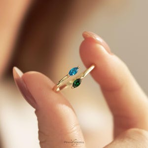 Könnte beinhalten: Ein goldener Bypass-Ring mit zwei marquise-geschliffenen Edelsteinen, einer in leuchtendem Blau und einer in Dunkelgrün, die an den offenen Enden des dünnen, polierten Goldrings sitzen. Der Text PinesPebbles ist unten im Bild zu sehen.