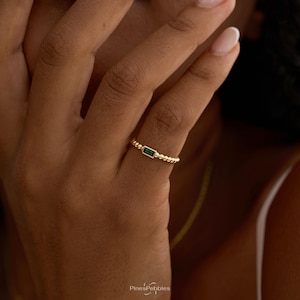 Könnte beinhalten: Ein goldener Ring mit einem kleinen, rechteckigen, smaragdgrünen Edelstein, der horizontal gefasst ist. Das Ringband ist mit einer feinen, perlenartigen Textur versehen, die den Finger umschließt. Der Text PinesPebbles ist unten sichtbar.
