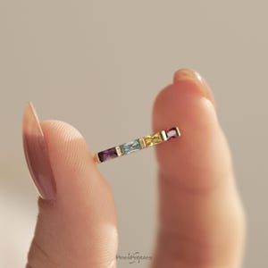 May include: A gold ring featuring four baguette-cut gemstones in a row, including a purple, light blue, yellow, and red stone. The band is thin and metallic, showcasing a multi-colored design with clear, vibrant gemstone colors.