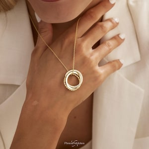 Peut inclure: Un collier doré avec un pendentif composé de trois anneaux entrelacés. Un anneau présente une texture perlée, tandis qu'un autre anneau lisse est gravé avec la date 15.08.26. Le pendentif est suspendu à une fine chaîne dorée.