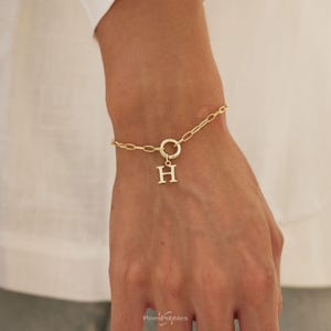 Könnte beinhalten: Ein goldfarbenes Kettenarmband mit ovalen Gliedern und einem runden Verschluss. Ein Anhänger in Form des Großbuchstabens H hängt in der Mitte des Verschlusses. Das Armband liegt um das Handgelenk und zeigt das metallische Finish und den Buchstaben.