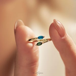 May include: A gold bypass ring featuring two marquise-cut gemstones, one in a vibrant blue and the other in a deep green. The open-ended band design allows for a slight adjustment, showcasing the contrasting colors of the two stones clearly.