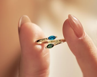 Puede incluir: Un anillo abierto de oro con dos piedras preciosas de talla marquesa, una en azul vibrante y la otra en verde intenso. El diseño de banda abierta permite un ligero ajuste, mostrando claramente los colores contrastantes de las piedras.