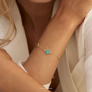 Könnte beinhalten: Ein goldenes Kettenarmband mit mehreren vierblättrigen Kleeblatt-Anhängern, die jeweils mit leuchtend türkisfarbenen Steinen besetzt sind. Die feinen Metallglieder verbinden die blütenförmigen Motive zu einem durchgehenden Muster.