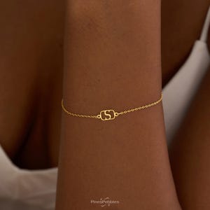 Könnte beinhalten: Ein zartes, goldfarbenes Kettenarmband mit einem zentralen rechteckigen Anhänger, der einen stilisierten Buchstaben S als Ausschnitt zeigt. Das Schmuckstück besteht aus einer feinen Gliederkette mit polierter Oberfläche und schlichtem Design.