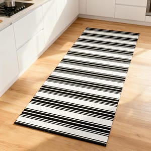 Puede incluir: Una alfombra rectangular de cocina con un patrón de rayas blancas y negras. La alfombra está colocada sobre un suelo de madera clara. Las rayas son horizontales y están espaciadas uniformemente, creando un aspecto clásico y moderno. La alfombra es larga y estrecha, adecuada para una cocina o pasillo.