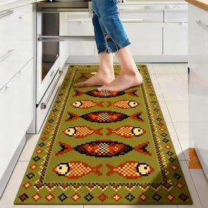 Op de afbeelding: Groene keukenmat met een pixelpatroon van vissen in oranje, zwart en bruin. De rechthoekige mat heeft een decoratieve rand. De mat ligt voor een keukenoven. De lengte van de mat is ongeveer 150 cm.