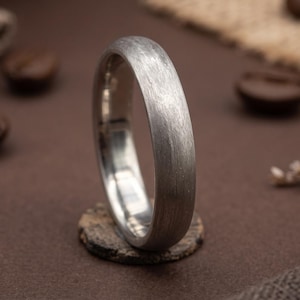 May include: A silver-toned ring with a brushed, matte finish and a rounded profile. The band features a textured surface that catches the light, showing a metallic sheen. The ring is positioned upright, displaying its circular shape and smooth interior.