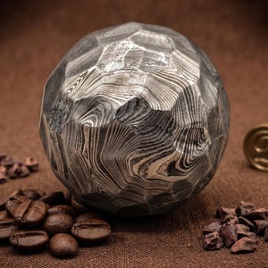 May include: A faceted sphere made of Damascus steel with a distinct layered, wavy pattern in shades of gray, black, and silver. The metal surface features a complex, swirling grain structure created by the forging process of the steel layers.