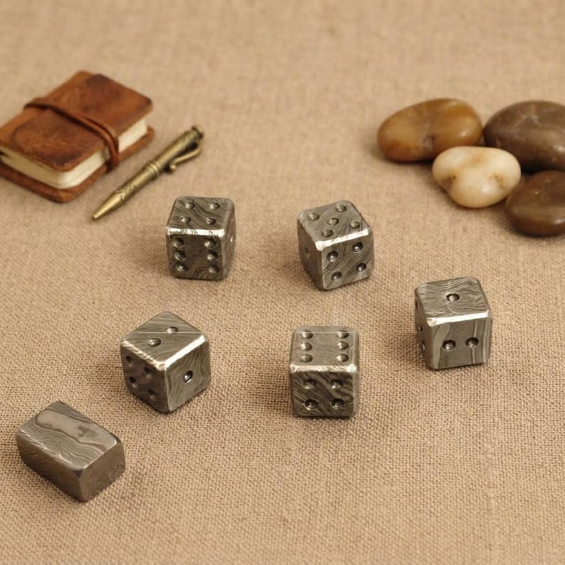 Pu&ograve; includere: Un set di sei dadi metallici caratterizzati da un distinto motivo a vortice grigio e argento, accompagnato da un piccolo taccuino rilegato in pelle marrone, una penna in metallo color oro e diversi ciottoli di fiume lisci e arrotondati.