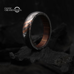 May include: A close-up of a ring with a patterned, dark metal exterior and a copper-colored accent. The interior of the ring is a warm brown wood. The ring is on a dark surface. The text "EXOTIC CREATIONDESIGN" is in the upper left corner.