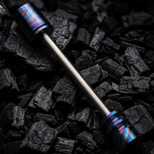May include: A metal tool with a cylindrical head and base, connected by a silver rod. The head and base have a vibrant, multicolored finish with hues of blue, purple, and orange. The tool rests on a bed of dark charcoal.