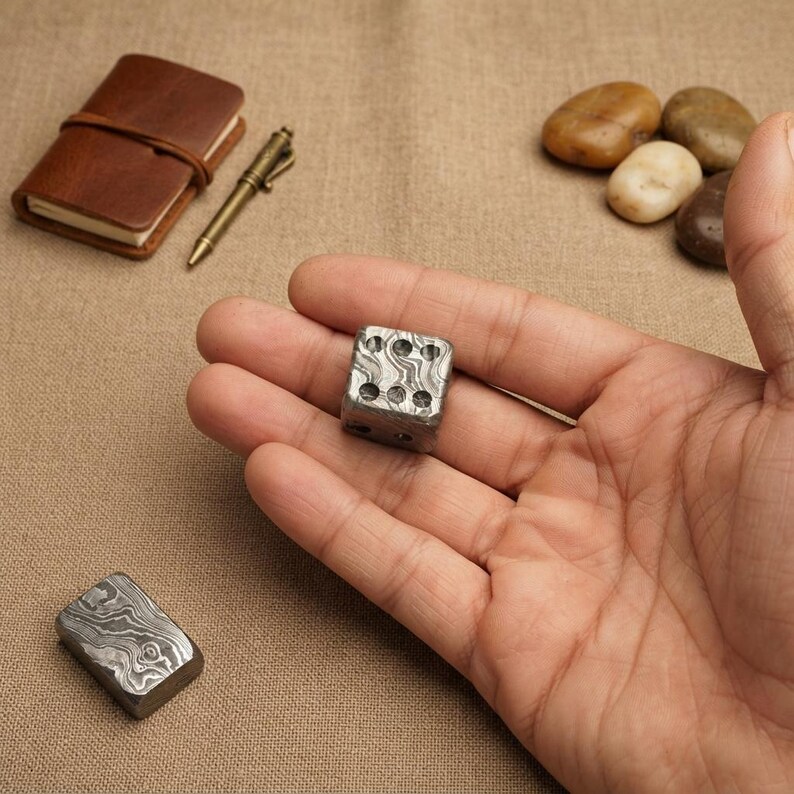 Pu&ograve; includere: Un dado a sei facce realizzato in acciaio Damasco con un distinto motivo metallico ondulato e stratificato. Il dado presenta rientranze circolari per i punti, mostrando una finitura grigio metallizzato e argento con una superficie lucida e testurizzata.