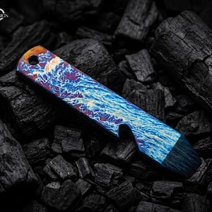 May include: A multi-colored bottle opener with a unique design. The opener features a vibrant pattern of blues, purples, and oranges, resembling a stormy sea. The opener is made of metal and has a hole at one end. The background is black.