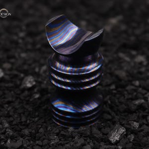 May include: A small, metallic bottle stopper with a unique design. The stopper has a ridged, cylindrical base and a curved top. The metal has a swirling pattern of blue, gold, and purple hues. The logo "EXOTIC CREATIONDESIGN" is in the top left corner.