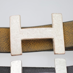 Pode incluir: Fivela de cinto de metal prateado em forma da letra "H" em um cinto de couro castanho. A fivela tem uma aparência texturizada e envelhecida. Um cinto de couro preto com uma fivela semelhante é parcialmente visível abaixo.