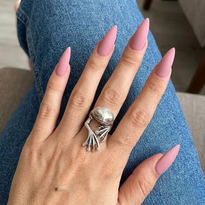 Puede incluir: Un anillo de metal plateado con forma de rana, usado en el dedo anular. El diseño de la rana presenta una cabeza lisa y redondeada con patas palmeadas detalladas que se extienden por el dedo. Las uñas son largas, puntiagudas y de color rosa mate.