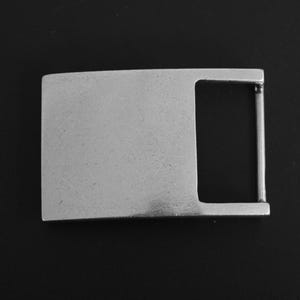 May include: A rectangular, silver-colored belt buckle with a cut-out section on the right side. The buckle has a brushed metal finish and a simple, minimalist design. The background is black.