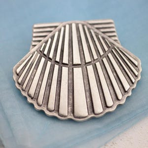 Op de afbeelding: Een zilverkleurig metalen schelpvormig object. Het ontwerp heeft een waaiervormig patroon van verticale lijnen die uitstralen vanuit een centraal punt. Het object heeft een rechthoekige basis en is waarschijnlijk een decoratief element of accessoire.