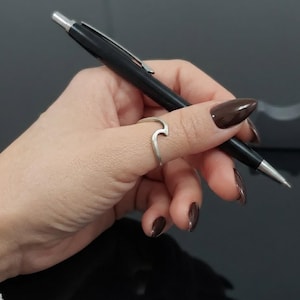 Könnte beinhalten: Ein silberner Ring mit einem wellenförmigen Designelement auf der Ringschiene. Der Ring wird an einem Finger getragen, und ein schwarzer Kugelschreiber mit silbernem Clip und Spitze wird in derselben Hand gehalten. Das Metall hat eine glatte Oberfläche.