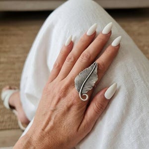 Può includere: Un anello color argento a forma di foglia stilizzata con una venatura centrale prominente e un gambo arricciato alla base. La superficie metallica presenta dettagli ossidati all'interno delle scanalature per evidenziare la trama del design.
